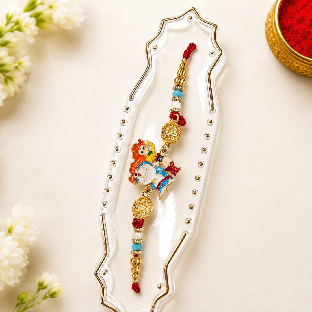A decorative Rakhi with a cartoon monkey charm, gold-tone coins, and red and blue beads, displayed on a white ceramic dish with white flowers and a gold bowl of red powder in the background.
