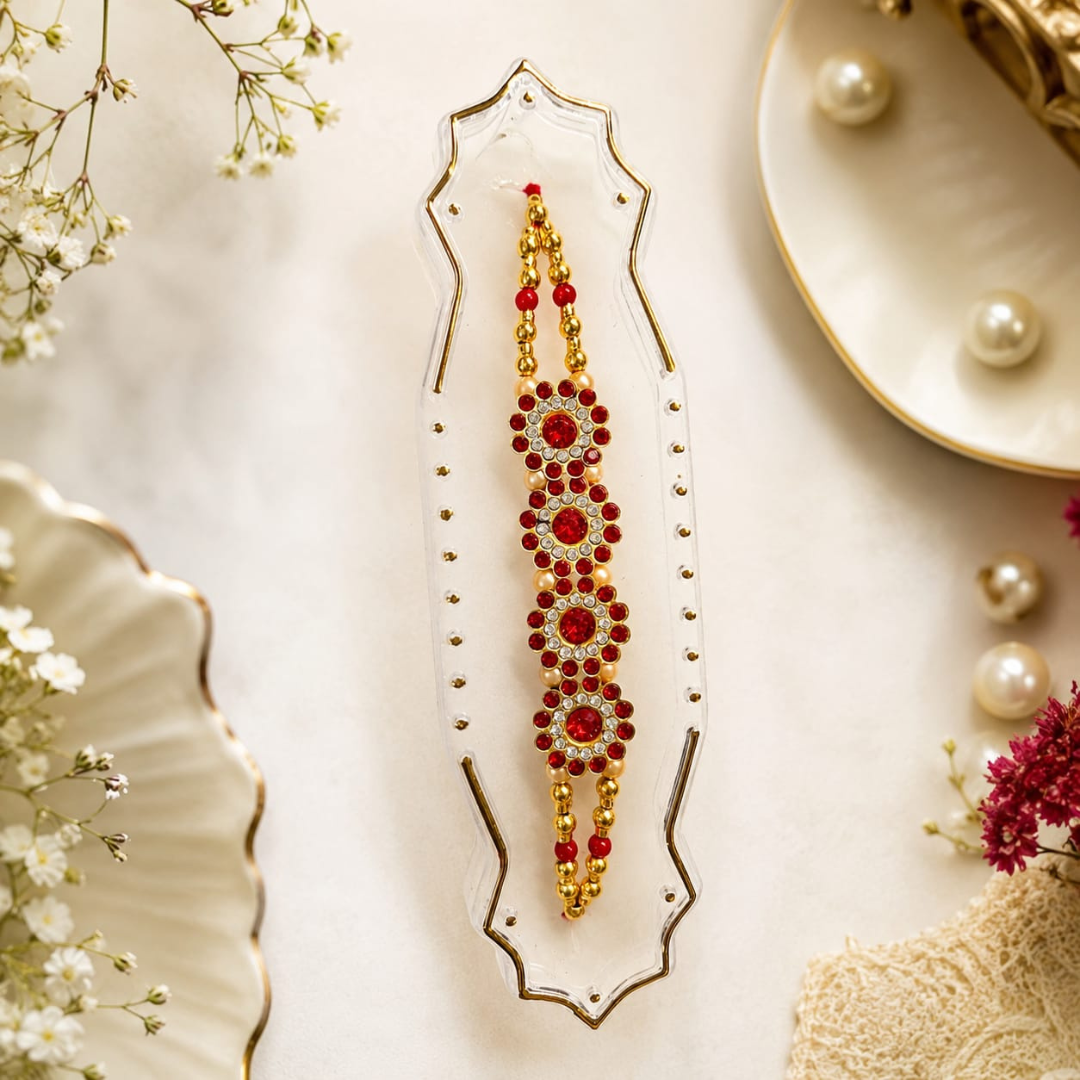 A gold and red beaded Rakhi bracelet displayed on a clear glass stand, surrounded by decorative floral elements and pearl accents on a light cream surface.