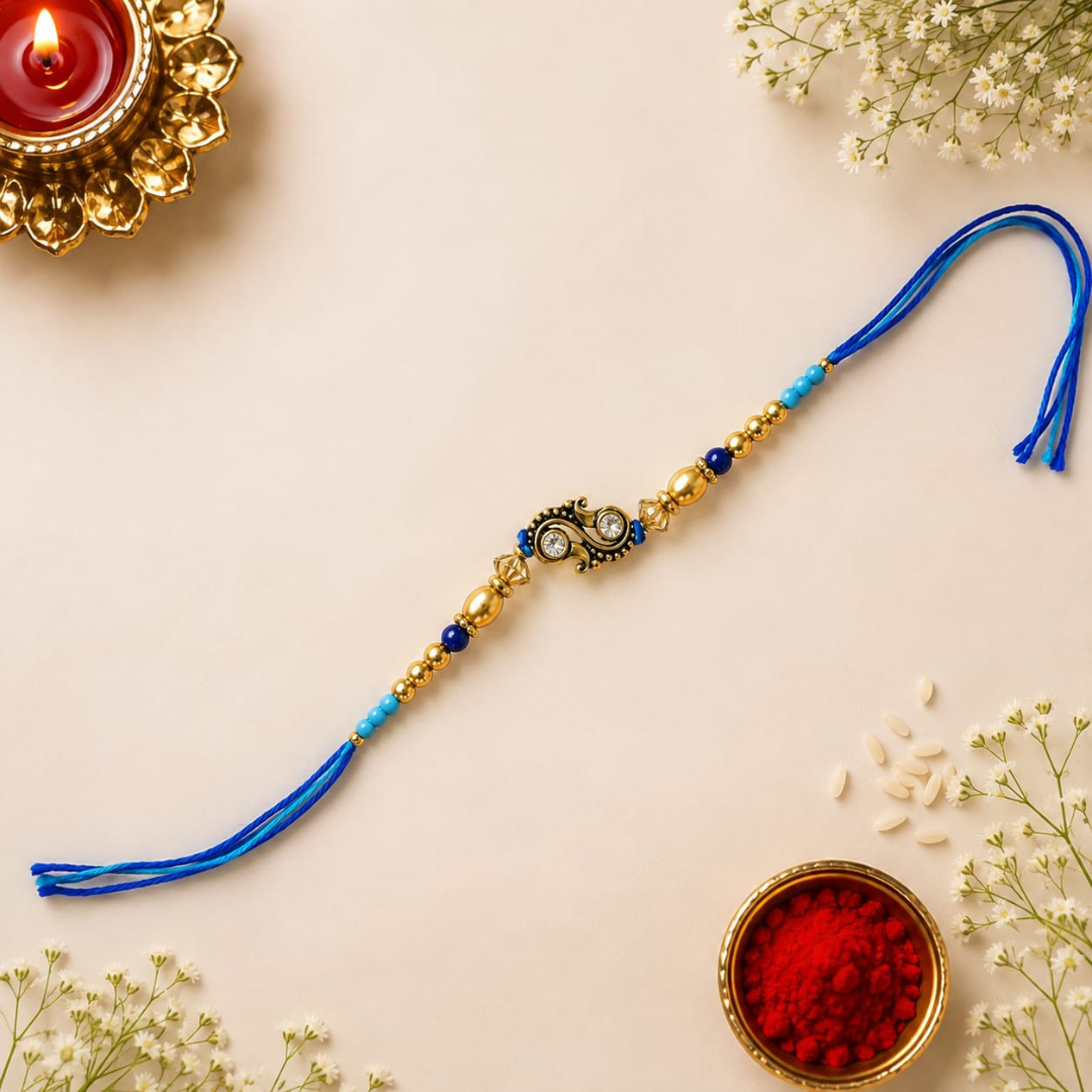 A traditional Indian Rakhi with blue thread tassels, gold beads, and a central decorative charm, laid out on a light beige background with festive props including a lit diya, rice, and red powder.