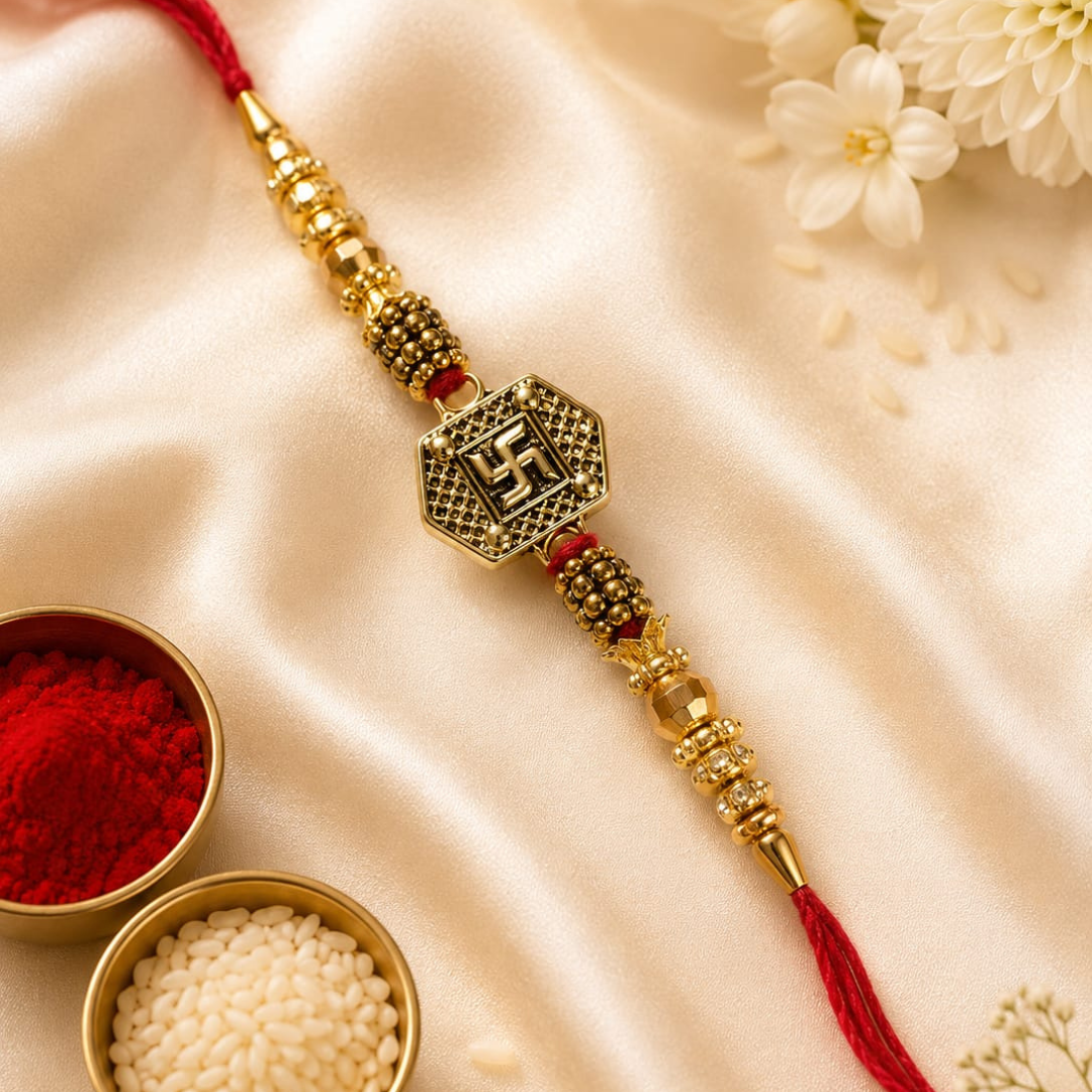 A gold-plated brass Lakshmi Rakhi with a central Swastika symbol charm, red cord, and red tassel, styled on a cream satin fabric with decorative white flowers and bowls of red powder and rice.