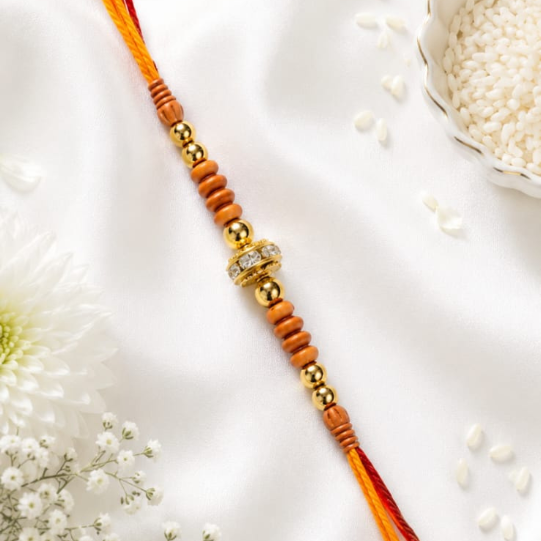 A traditional Indian Rakhi with orange and red cord, wooden beads, and gold-toned metal beads and spacer, laid diagonally on a white satin fabric with decorative white flowers and rice.