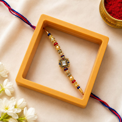 A decorative gold-toned Rakhi with red and blue thread accents and a central clear stone, displayed inside a mustard-yellow square box on a beige fabric surface with white flowers and a red powder bowl in the background.