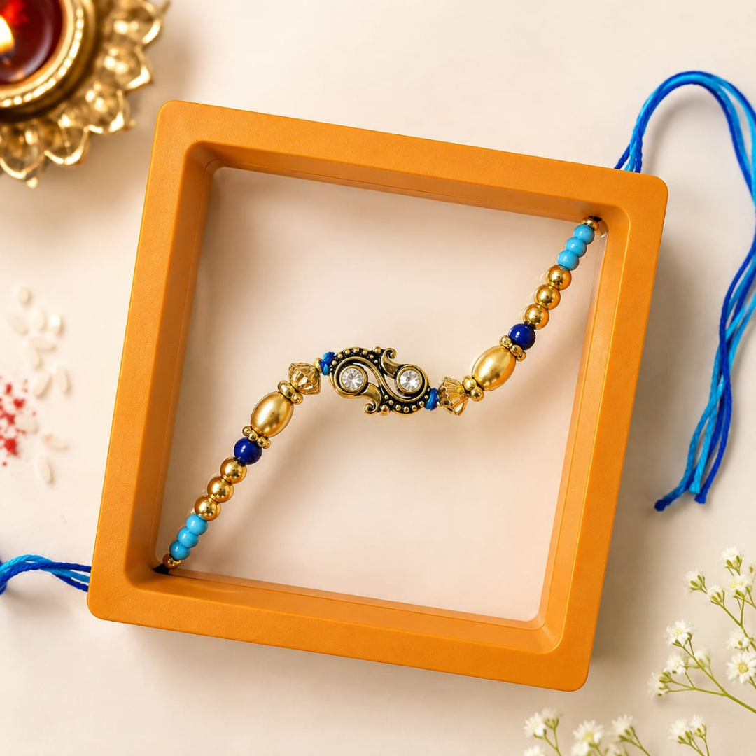 A decorative beaded bracelet with a central paisley motif, displayed inside an orange square box on a light beige surface with small white flowers and a brass lamp in the background.