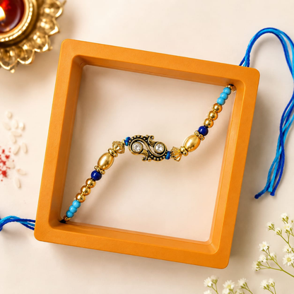 A decorative beaded bracelet with a central paisley motif, displayed inside an orange square box on a light beige surface with small white flowers and a brass lamp in the background.