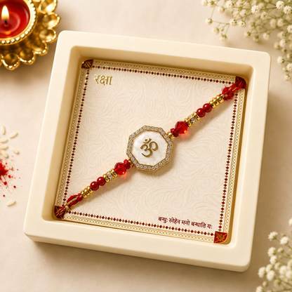 A red and gold beaded Rakhi with a central white and gold Om symbol, presented inside a cream-colored square gift box on a decorated festive background with a lit candle.
