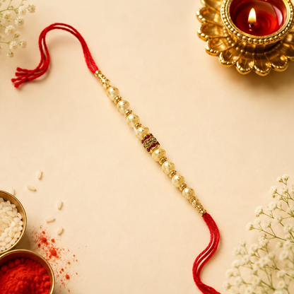 A traditional Indian beaded Rakhi with white pearl-like beads, gold spacer beads, and red tassels, laid diagonally on a beige surface with festive props including a lit diya, rice, red powder, and baby's breath.