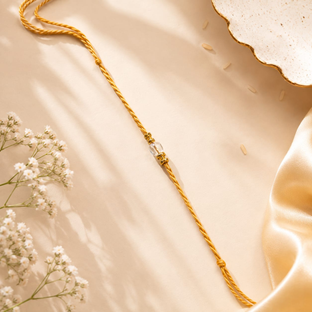 A gold-toned braided cord bracelet or necklace with a small clear faceted bead centerpiece, styled on a warm beige satin surface with white baby's breath flowers and a decorative dish edge.