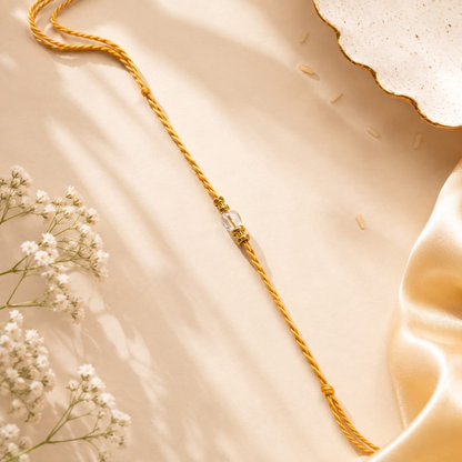 A gold-toned braided cord bracelet or necklace with a small clear faceted bead centerpiece, styled on a warm beige satin surface with white baby's breath flowers and a decorative dish edge.