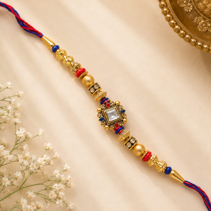 A traditional Indian gold-plated Rakhi bracelet with a central clear stone, red and blue thread accents, and gold beads, laid diagonally on a beige fabric background with small white flowers and a gold decorative plate in the corners.