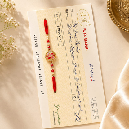 A traditional Indian Rakhi with red beads and a gold-toned circular pendant featuring a floral motif, laid flat on a cream-colored bank slip. The bank slip is styled on a beige surface with soft white flowers on the left and a gold decorative plate on the right.