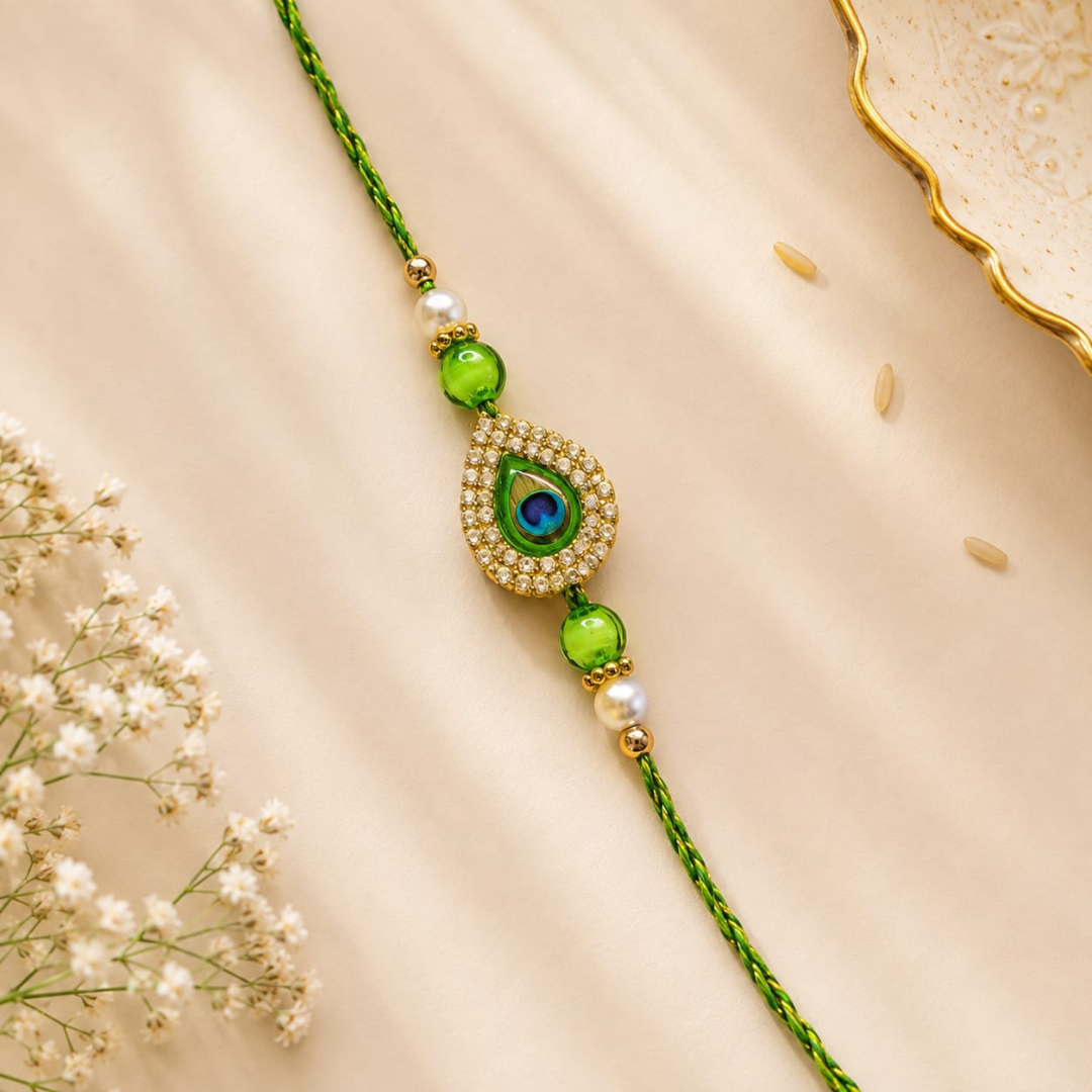 A green braided string bracelet with a central teardrop-shaped gold and crystal embellishment featuring a blue peacock motif, accented by green beads and white pearl-like beads, displayed on a beige satin fabric with baby's breath flowers.