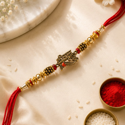A decorative Rakhi with a central metallic ornament, red tassels, and gold and black bead accents, laid diagonally on a beige satin fabric with small bowls of red powder and white rice nearby.