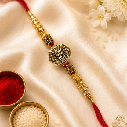 A gold-plated brass Lakshmi Rakhi with a central Swastika symbol charm, red cord, and red tassel, styled on a cream satin fabric with decorative white flowers and bowls of red powder and rice.