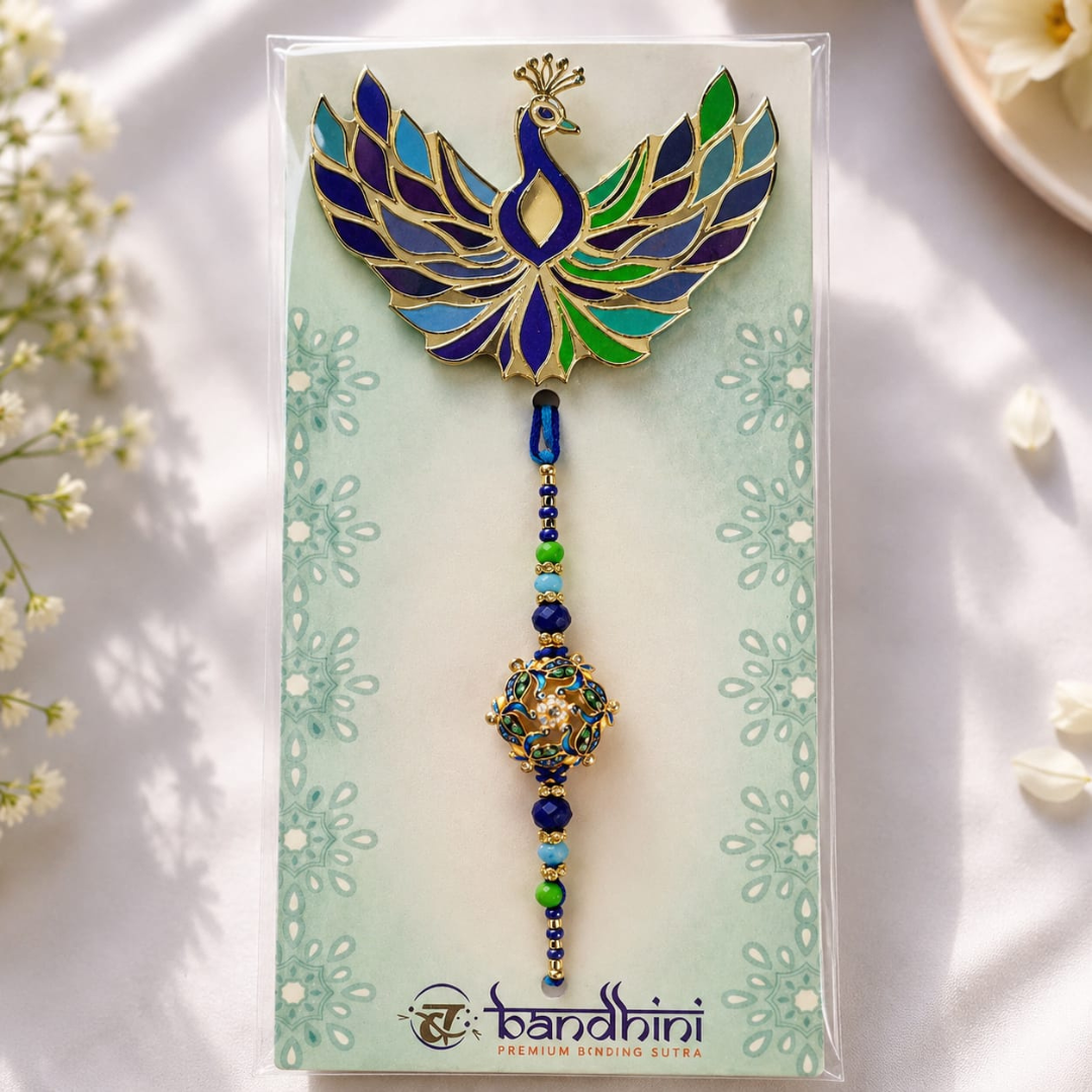 A decorative hanging ornament with a peacock-shaped top in blue, green, and gold, attached to a beaded tassel, displayed on a printed card. The card shows the brand name 'bandhini' and is photographed on a soft white surface with small floral props around it.