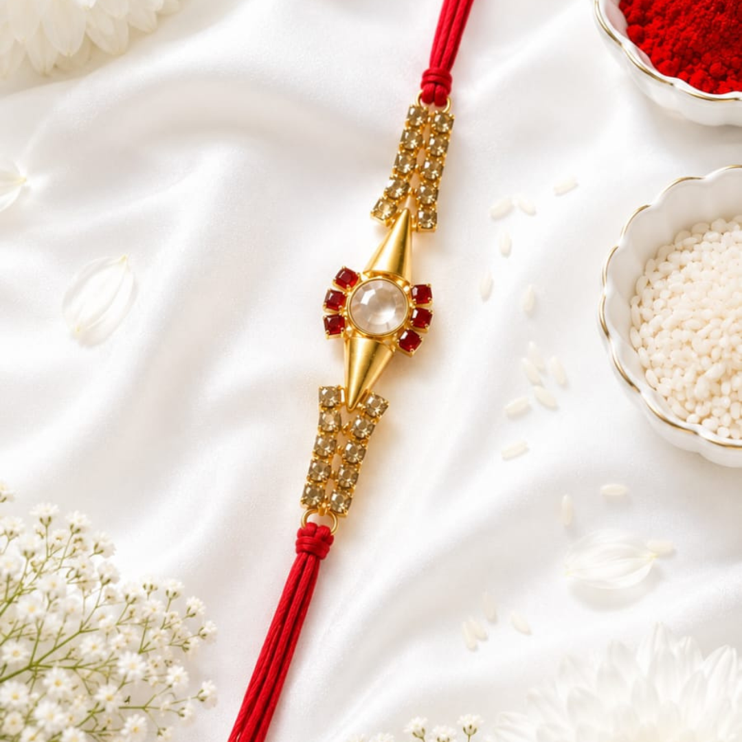 A gold and red Rakhi bracelet with a central white stone, laid diagonally on a white satin fabric with decorative white flowers and bowls of red powder and white grains nearby.