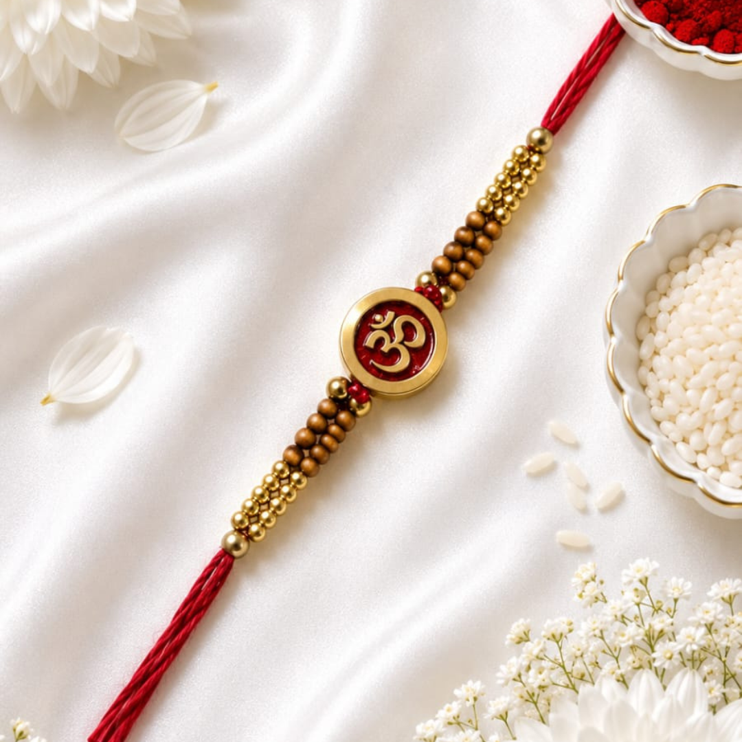 A red and gold beaded Rakhi with a central Om symbol charm, laid diagonally on a white satin fabric with decorative white flowers and bowls of rice.