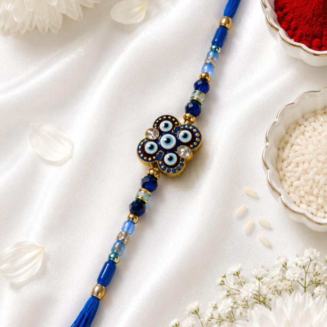 A blue and gold Rakhi with an evil eye charm, laid diagonally on a white satin fabric with decorative white flowers, a bowl of red powder, and a bowl of white rice.