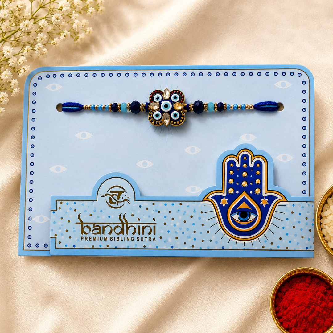 A blue and gold Hamsa-themed Rakhi bracelet presented on a branded light blue card, styled on a cream fabric background with small decorative flowers and bowls of red and white powder.