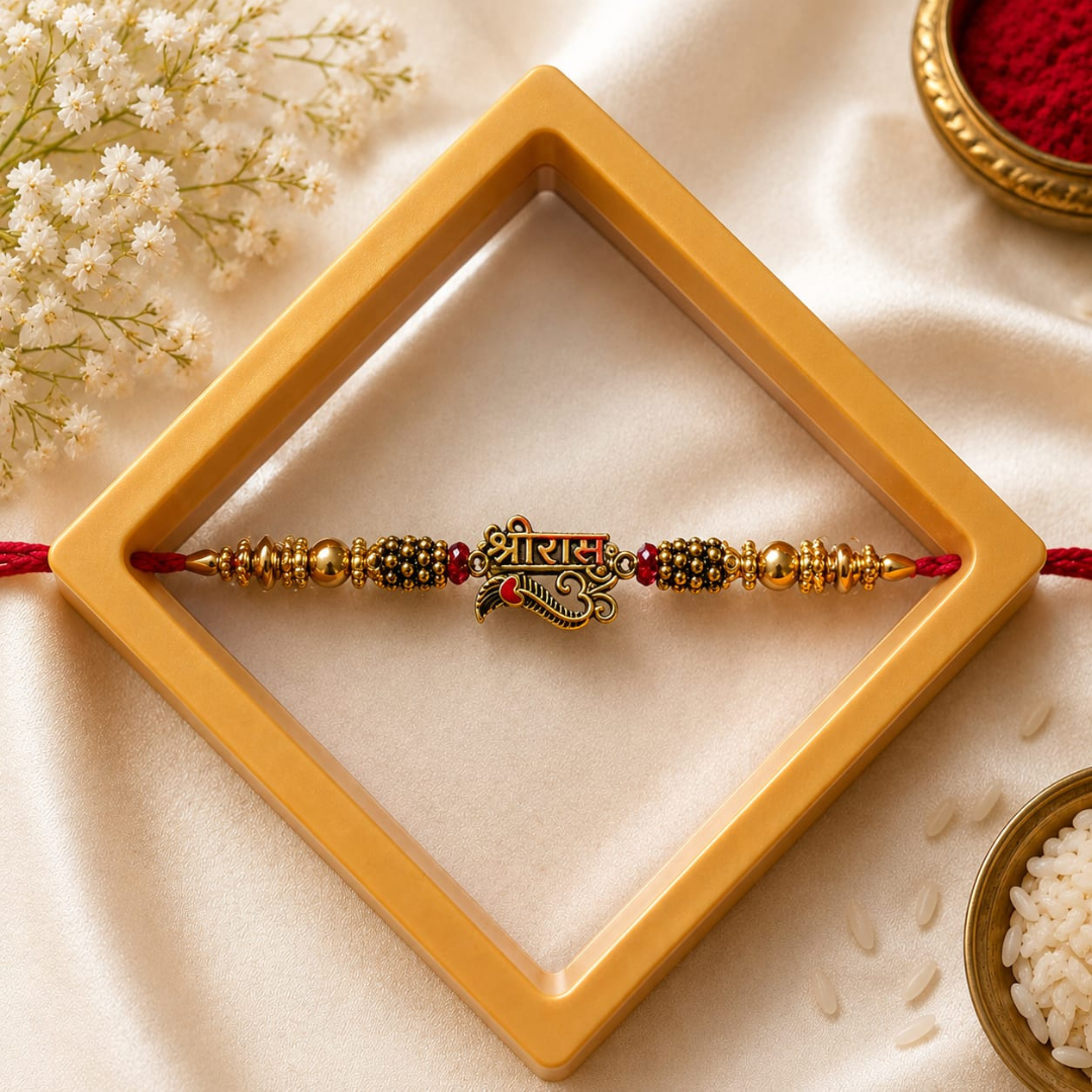 A gold and red beaded Rakhi with a central 'Mukut' ornament, presented inside a gold diamond-shaped frame on a cream satin fabric background with decorative flowers and rice.