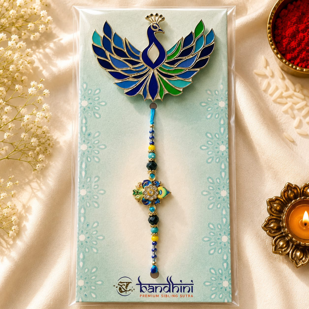 A blue and green peacock-shaped rakhi with colorful beaded strands, presented on a light blue decorative card by the brand bandhini, styled on cream fabric with flowers and a diya.